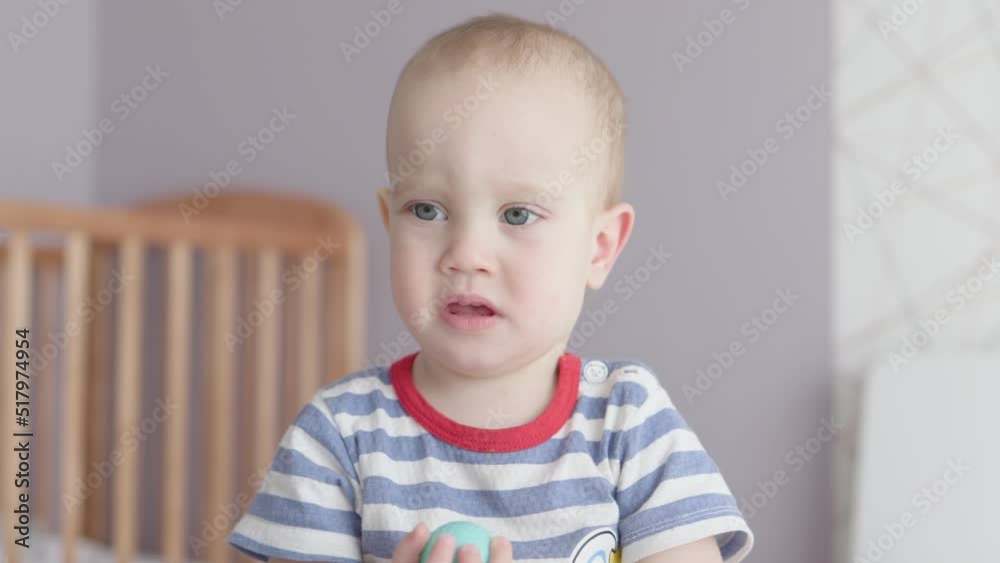 cute baby boy grimacing funny face child kid in striped blue red t-shirt in nursery room with brown cot crib in background. little baby bad mood starting to cry, depressed emotions, blue eyed toddler cute baby boy grimacing funny face child kid in striped blue red t-shirt in nursery room with brown cot crib in background. little baby bad mood starting to cry, depressed emotions, blue eyed toddler