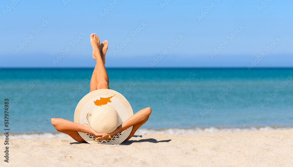 Fototapeta premium A slender tanned girl on the beach in a straw hat in the colors of the flag of Cyprus. Focus on the hat.