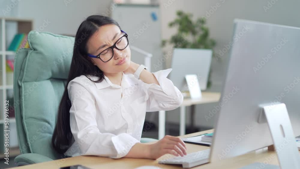 Tired Asian young female employee with pain in muscles of neck and spine from hard work sitting on workplace indoor. office worker woman feels ache discomfort massages suffering at computer desk. Asia