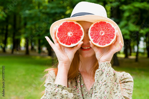 Summer portrait of young happy red haired female in dress and hat having fun in the park holding grapefruit. Vitamins,food.