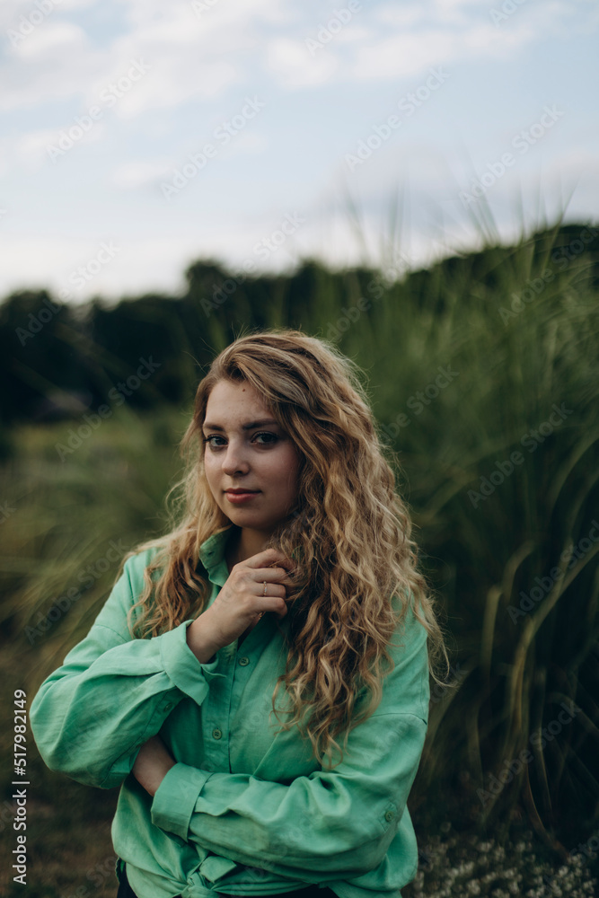 young beautiful woman with blond wavy hair posing near tall green grass. a plump girl in a green shirt and black trousers thought mysteriously about something.
