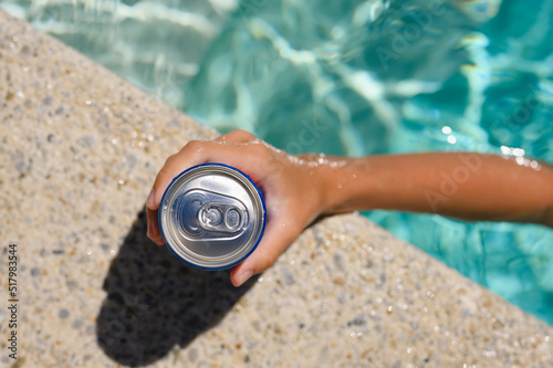 Can of cold drink in hand at the edge of the hotel pool. Top view, selective focus on the bank.