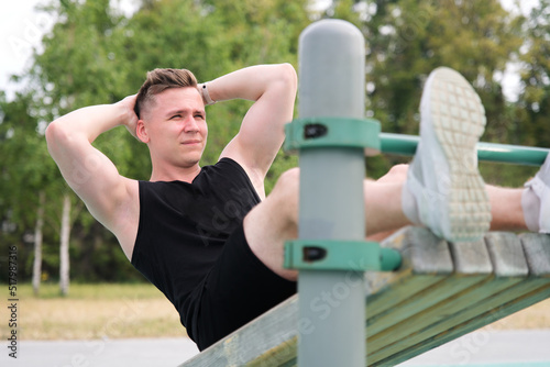 Happy fit fitness handsome athletic guy, young man training abs on bench, doing abdominal exercise on a public equipment in outdoor fitness park. Workout and bodybuilding concept, healthy lifestyle.