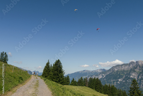 Three hikers hiking a path in the Flumserberg, in Walenstadt, near Zurich, Switzerland, while paragliders flying on a blue sky summer day.