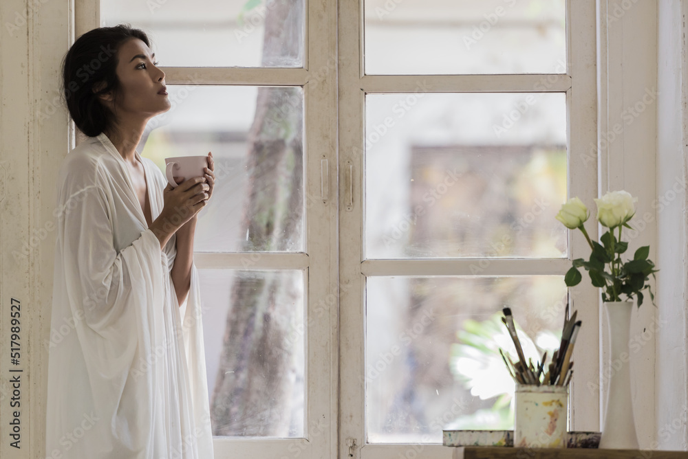 Happy young woman at home standing near window relaxing in her living room and drinking a cup of coffee. Happy young woman at home standing near window relaxing in her living room and drinking a cup of coffee.