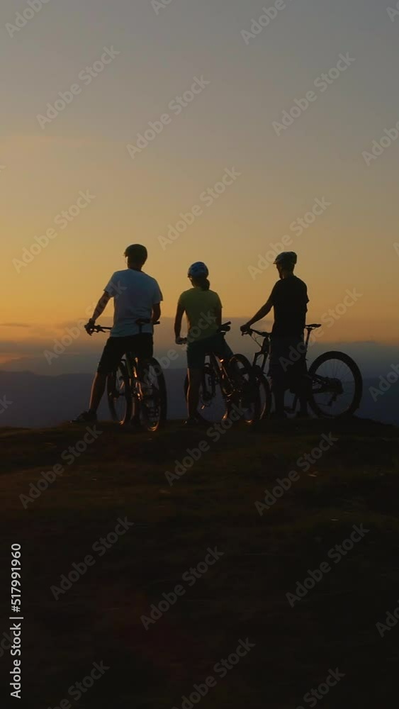 DRONE, SILHOUETTE, LENS FLARE: Mountain bikers stop and observe the sunset after an adrenaline filled downhill adventure. Three friends watch the sunset after a bicycle ride in the scenic mountains
