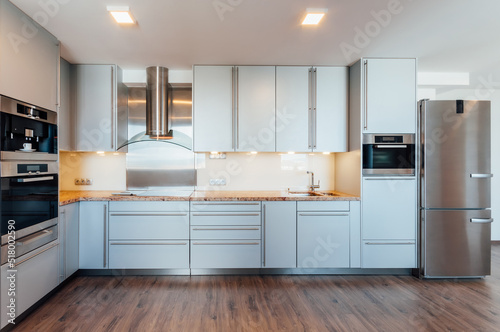 Large luxury kitchen unit made of light blue laminate, glass and marble. It is equipped with premium built-in appliances. The kitchen is lit by warm spot LED lights. The floor is wooden.