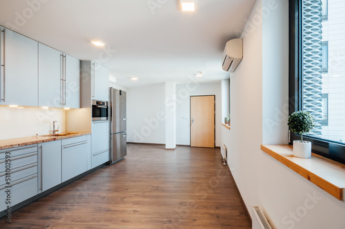 Room in apartment with visible part of luxury kitchen unit with sink  microwave and refrigerator. There is door on back wall and two windows to right  between which the ac unit is located on the wall.