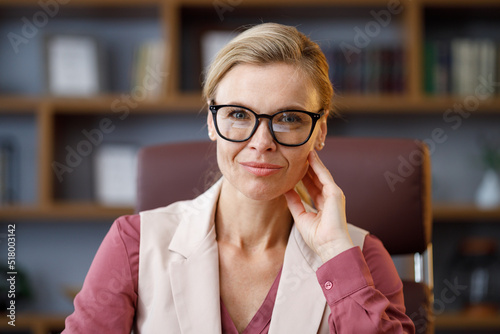 Portrait of beautiful blondie woman looking at camera and smiling. Adult stylish confident businesswoman wearing eyeglasses sitting at workplace in office. Portrait of modern successful female leader