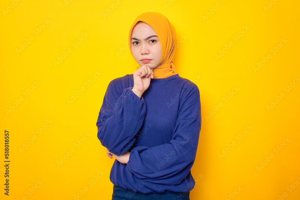 Pensive young Asian Muslim woman dressed in casual sweater holding hand on chin and looking at camera isolated over yellow background Pensive young Asian Muslim woman dressed in casual sweater holding hand on chin and looking at camera isolated over yellow background