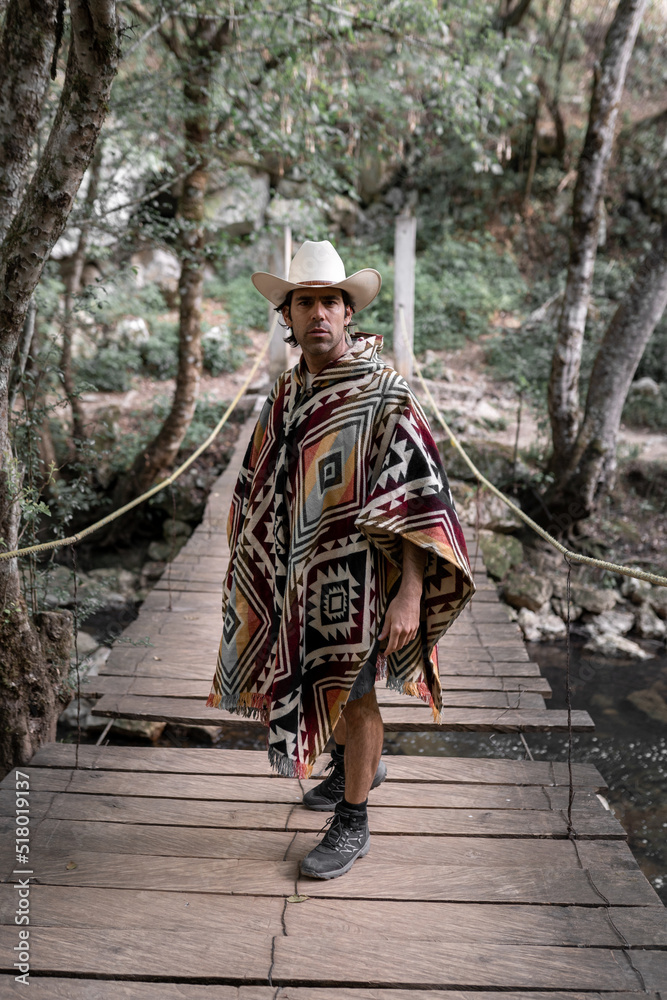 A mexican young man wearing a poncho and a hat is posing on a wooden bridge