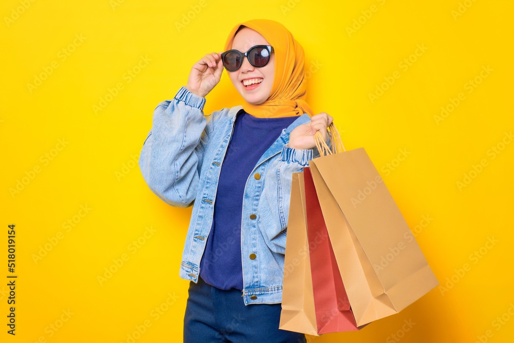 Happy young Asian woman in jeans jacket holding shopping bags and touching sunglasses, looking at camera isolated over yellow background