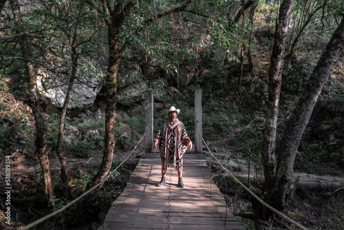Mexican young man wearing a poncho and a hat is standing with a duel pose on a wooden bridge