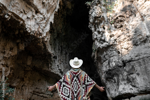 Unrecognizable mexican man wearing a poncho and a hat is standing in front of a cave with open arms