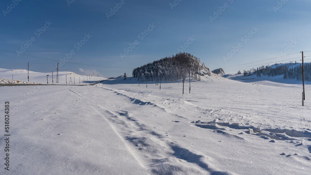 Tire tracks are visible in the endless snow-covered valley. The road goes to the horizon. Bare trees and mountains against the blue sky. Altai. Chuysky tract