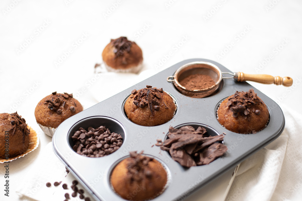 Chocolate muffins flat lay in baking tray with slides of chocolate, chocolate chip and cocoa powder on white cutting board and white cloth in soft focus Chocolate muffins flat lay in baking tray with slides of chocolate, chocolate chip and cocoa powder on white cutting board and white cloth in soft focus
