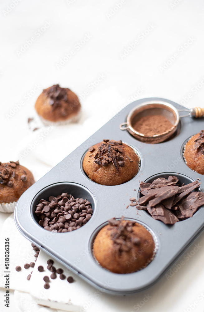 Chocolate muffins flat lay in baking tray with slides of chocolate, chocolate chip and cocoa powder on white cutting board and white cloth in soft focus