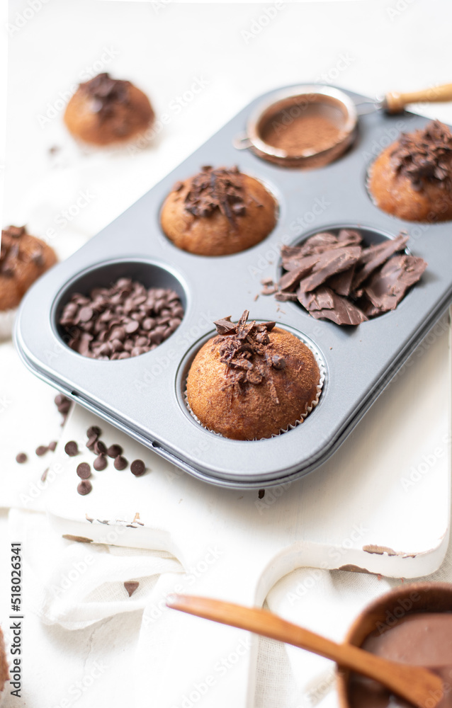 Chocolate muffins flat lay in baking tray with slides of chocolate, chocolate chip, cocoa powder and chocolate sauce on white cutting board and white cloth in soft focus Chocolate muffins flat lay in baking tray with slides of chocolate, chocolate chip, cocoa powder and chocolate sauce on white cutting board and white cloth in soft focus