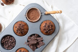 Close up top view of chocolate muffins flat lay in baking tray with slides of chocolate, chocolate chip and cocoa powder on white cutting board and white cloth