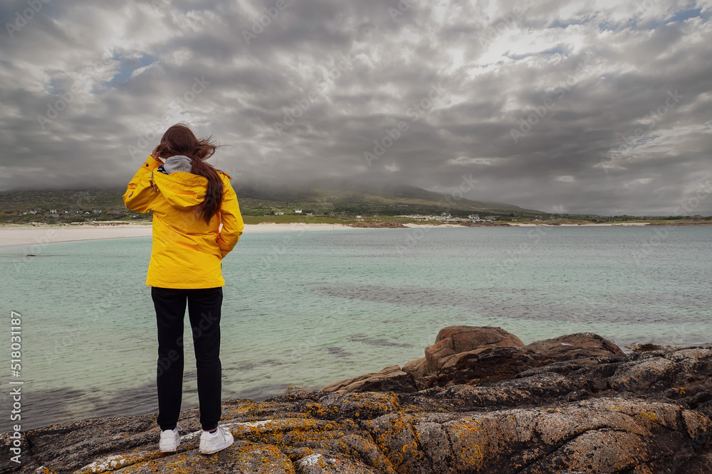 Teenager girl standing on a rock, looking at amazing Gurteen beach, county Galway, Ireland in the background. Popular tourism area with amazing nature scene. Irish landscape. Travel concept
