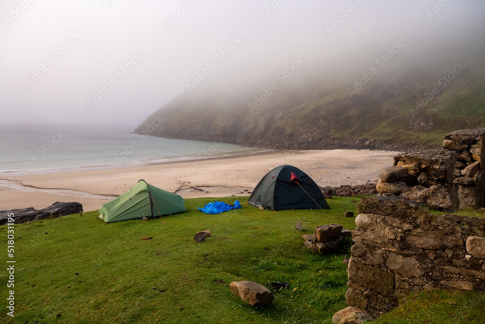 Two tents set up on a green grass patch of land. Mountain and sandy beach and ocean in the background. Morning fog over water. Keem beach, Ireland. Travel and tourism concept. Campsite in nature area.