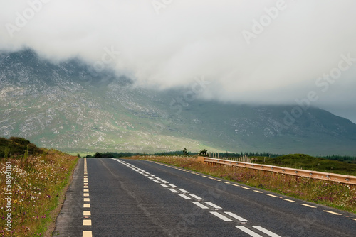 Straight asphalt road to a mountain covered with low clouds. Transportation and tourism. West of Ireland. Calm and peaceful mood. © mark_gusev