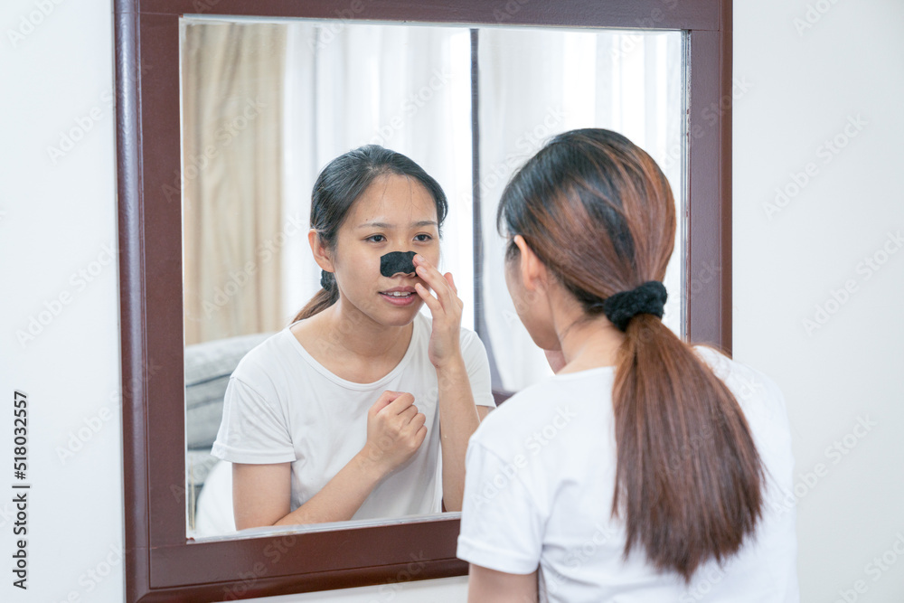 Beautiful Asian woman looking at the mirror, applying mask charcoal deep cleansing nose pore strip on fresh clean skin. Beauty and skin care treatment concept. Beautiful Asian woman looking at the mirror, applying mask charcoal deep cleansing nose pore strip on fresh clean skin. Beauty and skin care treatment concept.