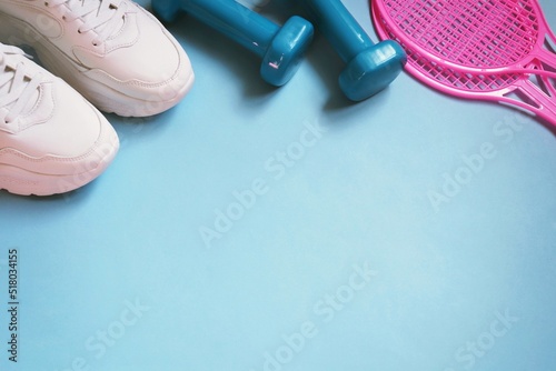 Sports equipment photo top view. White sneakers, dumbbells and pink tennis rackets on a blue background. Fitness mockup for banner design © learesphoto