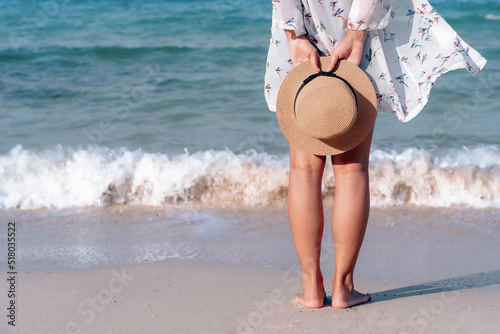 Back view of woman's legs standing on sandy beach near turquoise colored ocean in summertime. Female tourist with straw hat in hands enjoying traveling to exotic nature on a beautiful sunny day.
