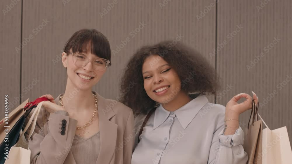 Medium slowmo of two young beautiful diverse girlfriends with many paper shopping bags smiling at camera after shopping for clothes in shopping center Medium slowmo of two young beautiful diverse girlfriends with many paper shopping bags smiling at camera after shopping for clothes in shopping center