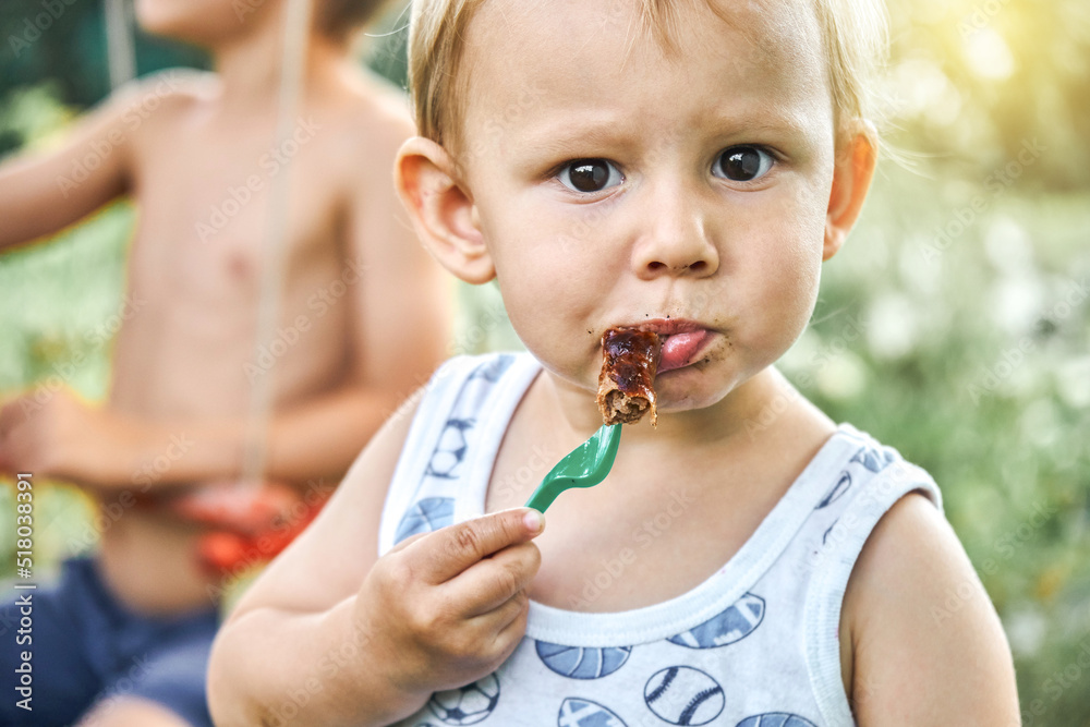 Cute blond toddler boy eats barbecue sausage on blurred background Cute blond toddler boy eats barbecue sausage on blurred background