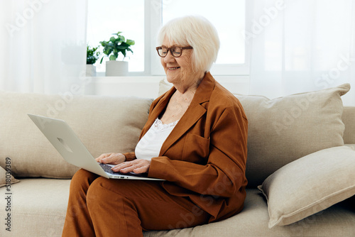 a sweet, enthusiastic elderly lady with gray hair is sitting on the couch working remotely on a laptop and smiling with joy enjoying the process