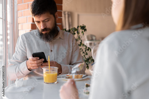 Cheerful spouses using smartphone while eating breakfast in kitchen  happy couple enjoying tasty morning meal  shopping online or checking social networks on cellphone
