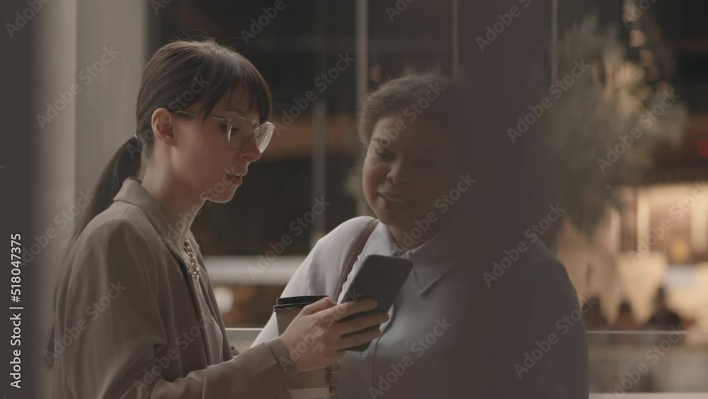 Waist up slowmo of two young multiethnic female colleagues looking at smartphone and chatting at modern shopping or business center Waist up slowmo of two young multiethnic female colleagues looking at smartphone and chatting at modern shopping or business center