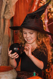 Smiling red-haired girl in a black witch costume and hat in Halloween decorations.Halloween concept.Selective focus.