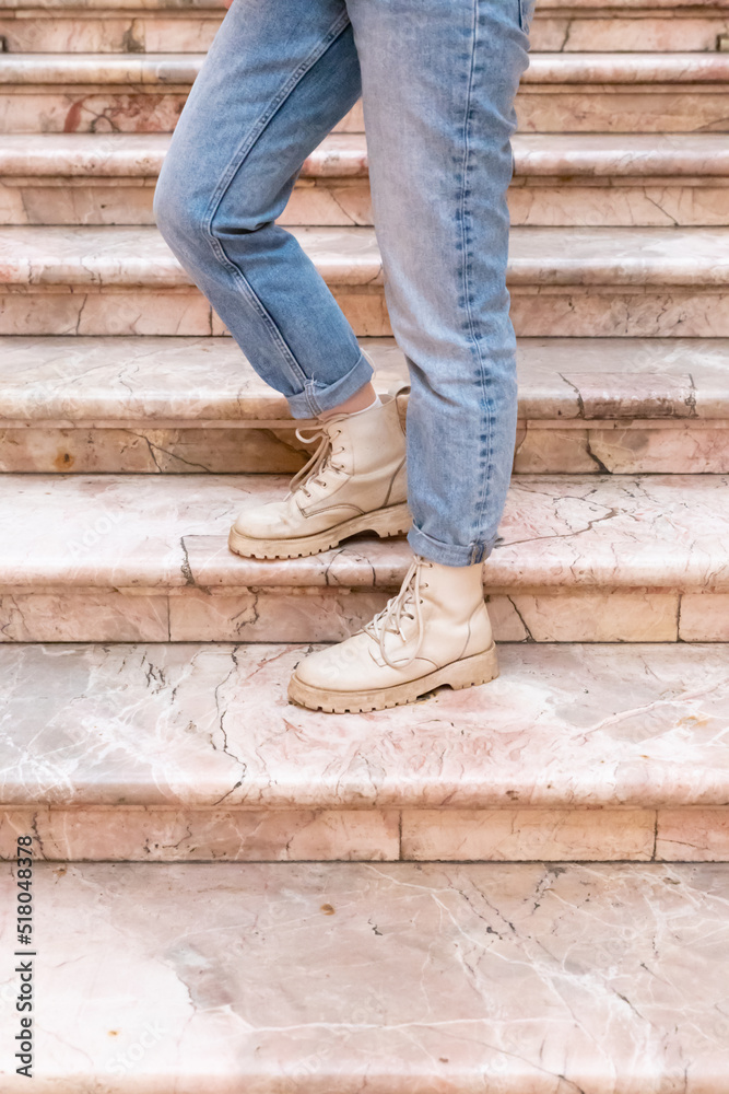 Woman's legs in blue twisted jeans and in beige boots on pink marble staircase. Front view, vertical photo