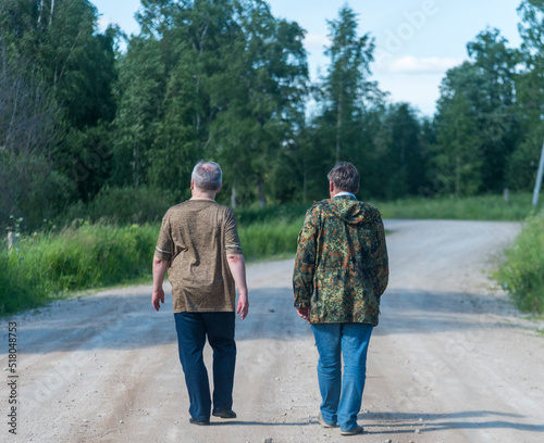 two men are walking away along a rural dusty country road among the trees © Dmitry