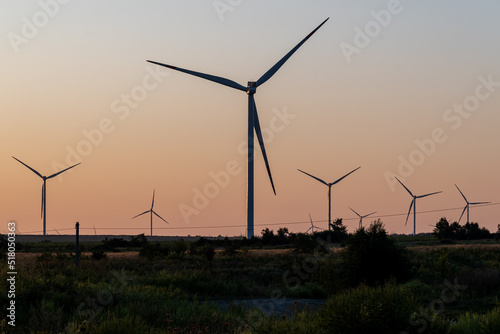 Large ecological windmills in endless green field against backdrop of setting sun.