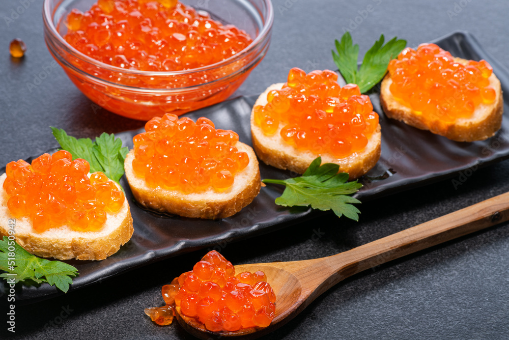 Red caviar on small buns with small ones that lie on black plate on wooden spoon and in glass plate on black background. Caviar on spoon on buns and in plate. Red caviar on small buns with small ones that lie on black plate on wooden spoon and in glass plate on black background. Caviar on spoon on buns and in plate.