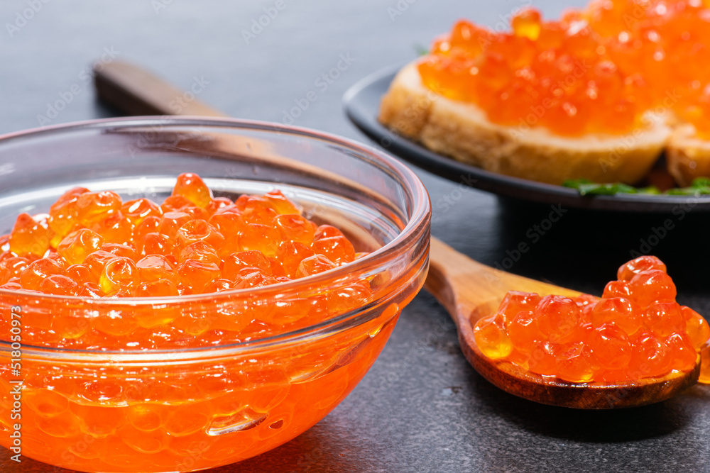 Red caviar in large glass and transparent plate, behind is wooden spoon with caviar and white small buns on black plate on gray background. Red caviar in large glass and transparent plate, behind is wooden spoon with caviar and white small buns on black plate on gray background.