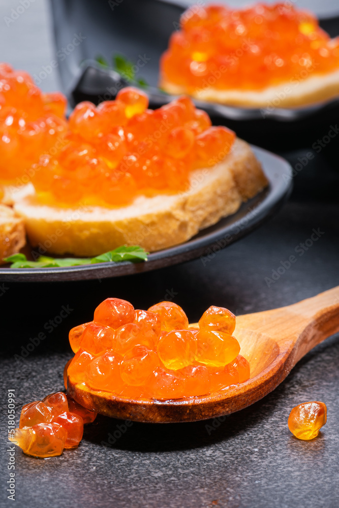 Red caviar in wooden spoon and on rolls with butter and green parsley on black plates on gray table. red caviar close-up in spoon and on butter rolls. Red caviar in wooden spoon and on rolls with butter and green parsley on black plates on gray table. red caviar close-up in spoon and on butter rolls.