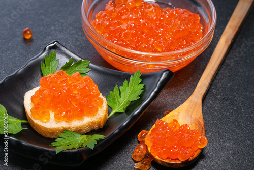 Red caviar on bun with small one, in small wooden spoon and glass plate on black background. Caviar on black background.