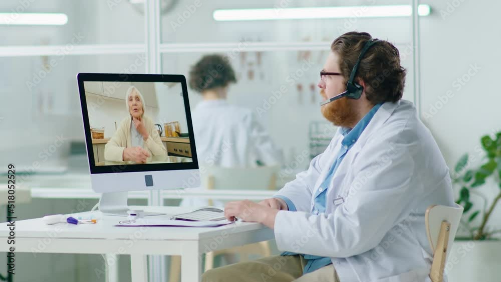 Male doctor in headset talking with eldelry woman via online video call on computer while giving online health consultation from medical office