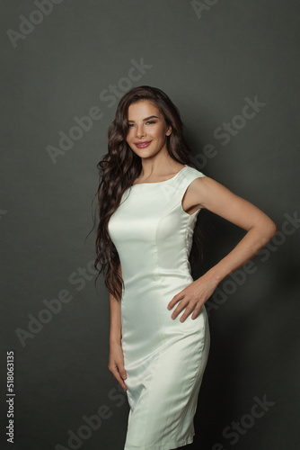 Positive brunette woman with long dark hair smiling on black background. Fashion model wearing white silky gown © millaf