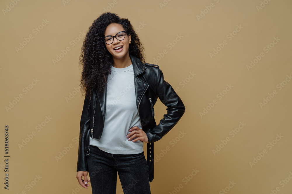 Fashionable woman with glad expression, keeps hand on hip, smiles cheerfully, wears optical glasses, white t shirt, black leather jacket and jeans, ready for walk, poses against beige background Fashionable woman with glad expression, keeps hand on hip, smiles cheerfully, wears optical glasses, white t shirt, black leather jacket and jeans, ready for walk, poses against beige background