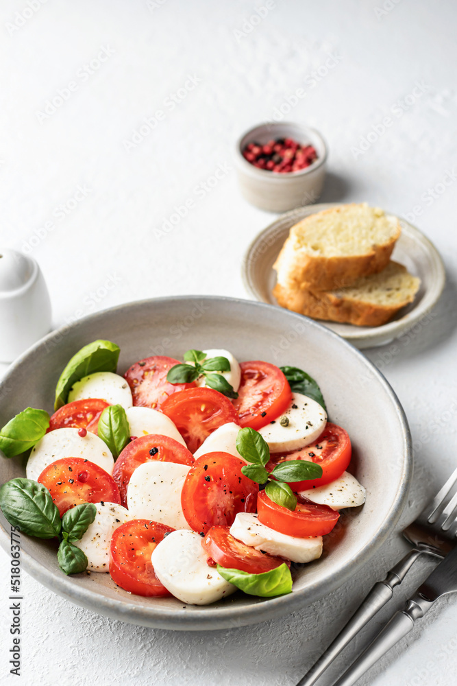 Caprese salad with tomatoes mozzarella basil and olive oil on white background served with cutlery and bread. Text space