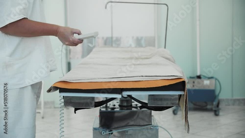 Close-up of scrub nurse pressing buttons on remote control, modern automatic motorized surgical operating table rotates and changes tilt angle, hi-tech equipment in hospital surgery operating room.
