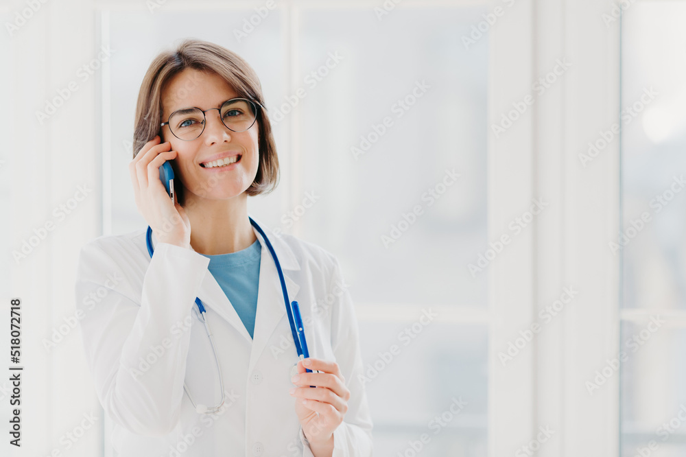 Positive woman doctor consults patient via cellphone, looks gladfully at camera, holds pen, wears white coat, poses in medical office, poses near window. Healthcare and consultation concept. Positive woman doctor consults patient via cellphone, looks gladfully at camera, holds pen, wears white coat, poses in medical office, poses near window. Healthcare and consultation concept.