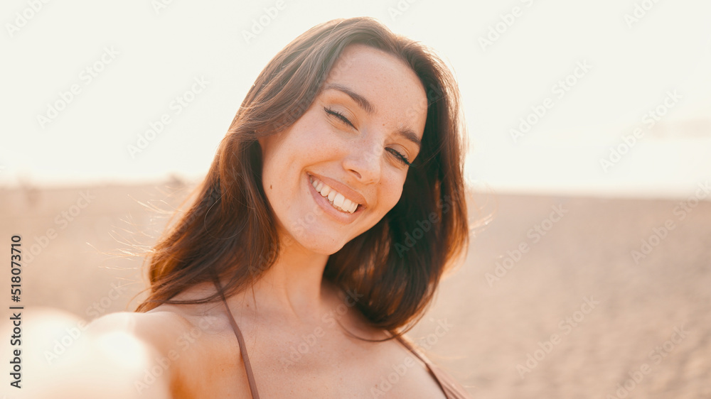 Clouse-up, beautiful brown-haired woman with long hair uses mobile phone. Smiling girl wearing swimsuit takes selfie on the beach on seascape background.