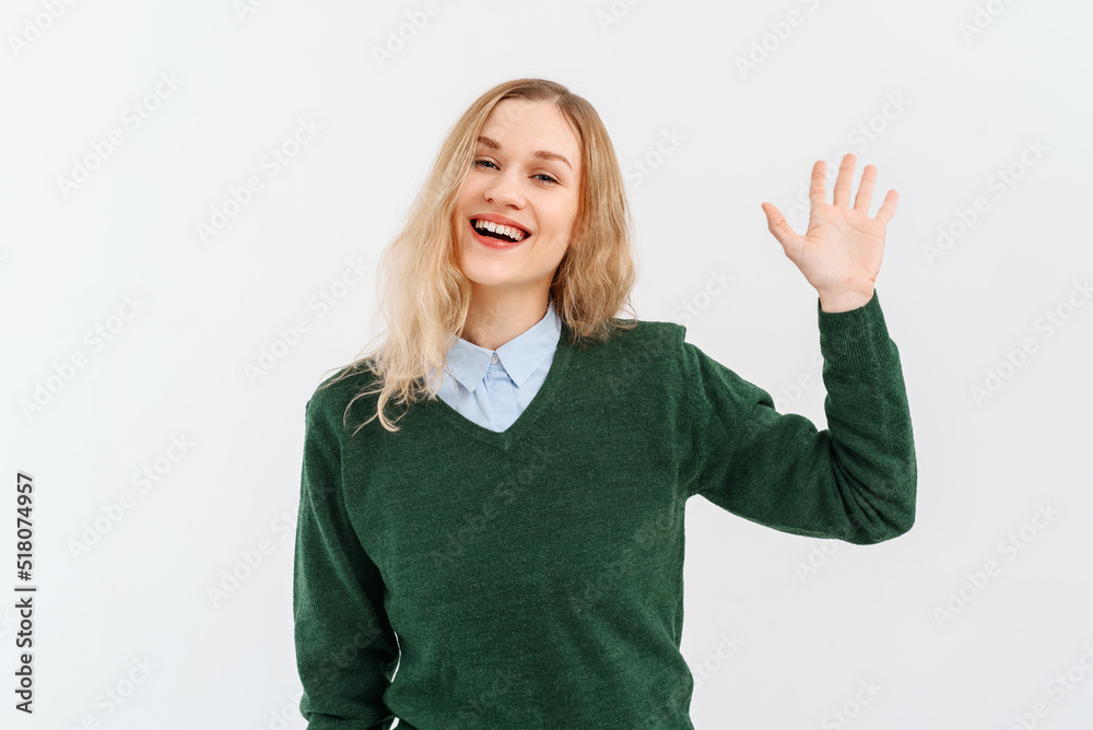 Friendly cheerful blonde girl smiling, saying hi and waving hand to greet person, make hello gesture, welcome someone, wears casual clothes. Indoor studio shot on white background